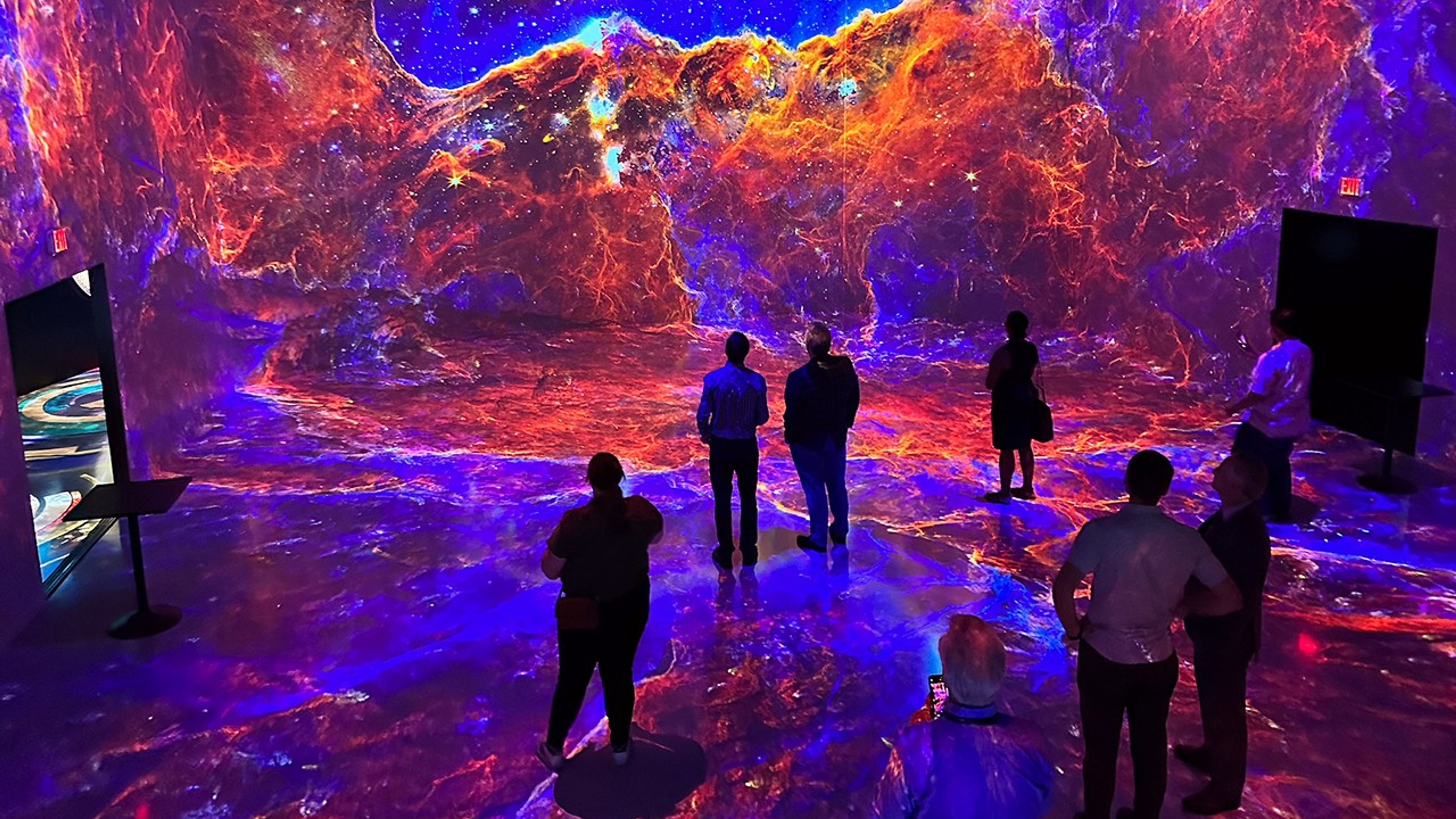 Images incorporating science data from the James Webb Space Telescope are projected onto the walls of a large room as part of a digital art piece. Seven guests stand in the room watching the show.