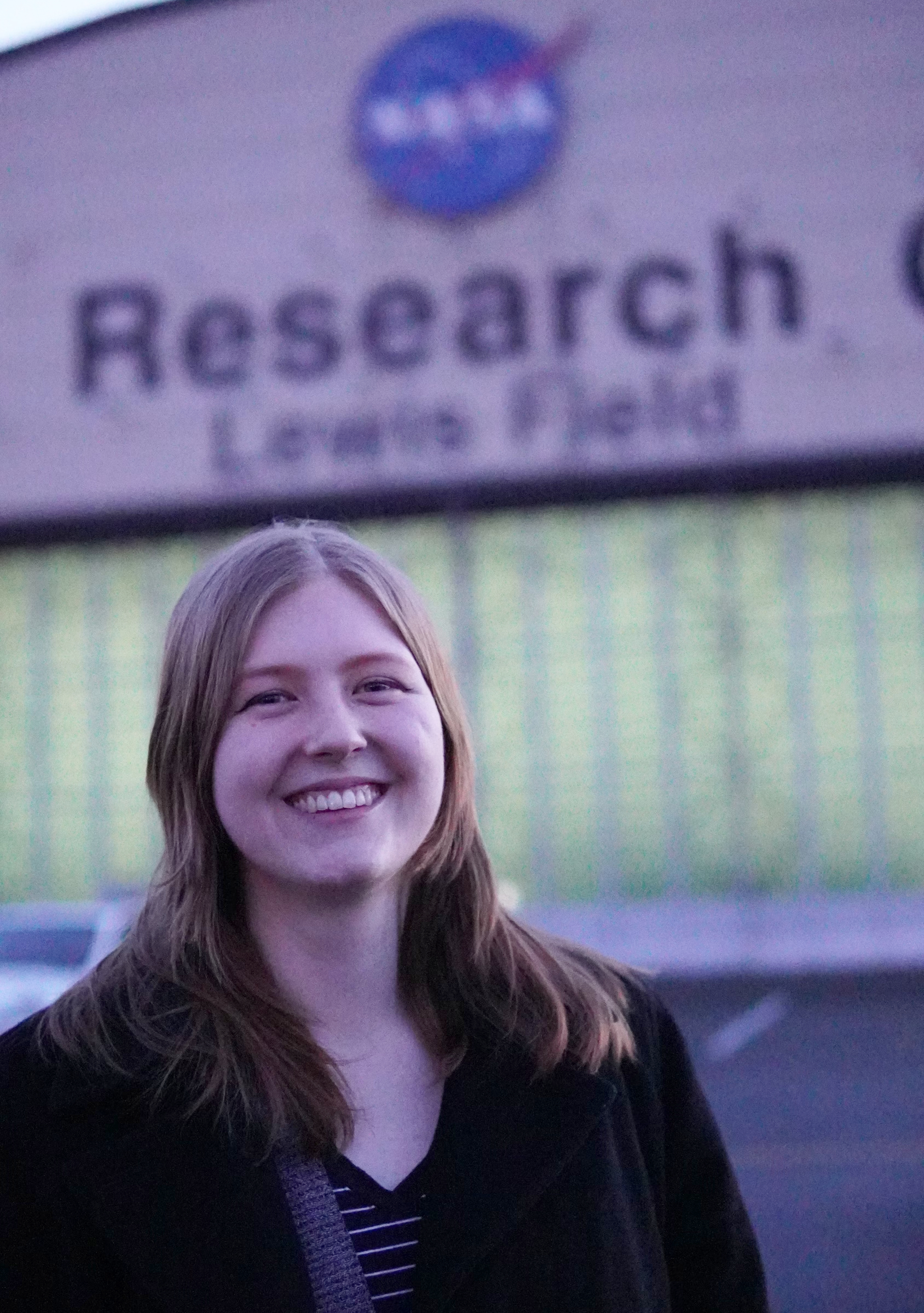 A headshot of Lauren Leese, a white woman with long reddish-blond hair wearing a black coat, standing in front of the main hangar at the NASA Glenn Research Center in Cleveland.