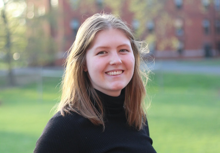 A headshot of Lauren Leese, a white woman with long reddish-blond hair wearing a black sweater, standing in front of a lawn.